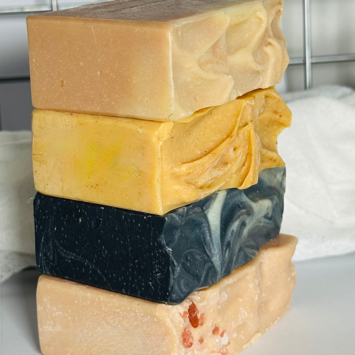 A stacked set of 4 artisanal handmade soaps in various colours and textures, including marbled black and white, yellow swirls, and solid tones, photographed close-up.
