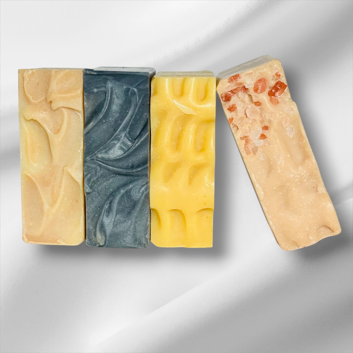 Four artisanal handmade soap bars arranged on a white draped fabric background. The soaps feature textured swirls and embedded botanical elements.