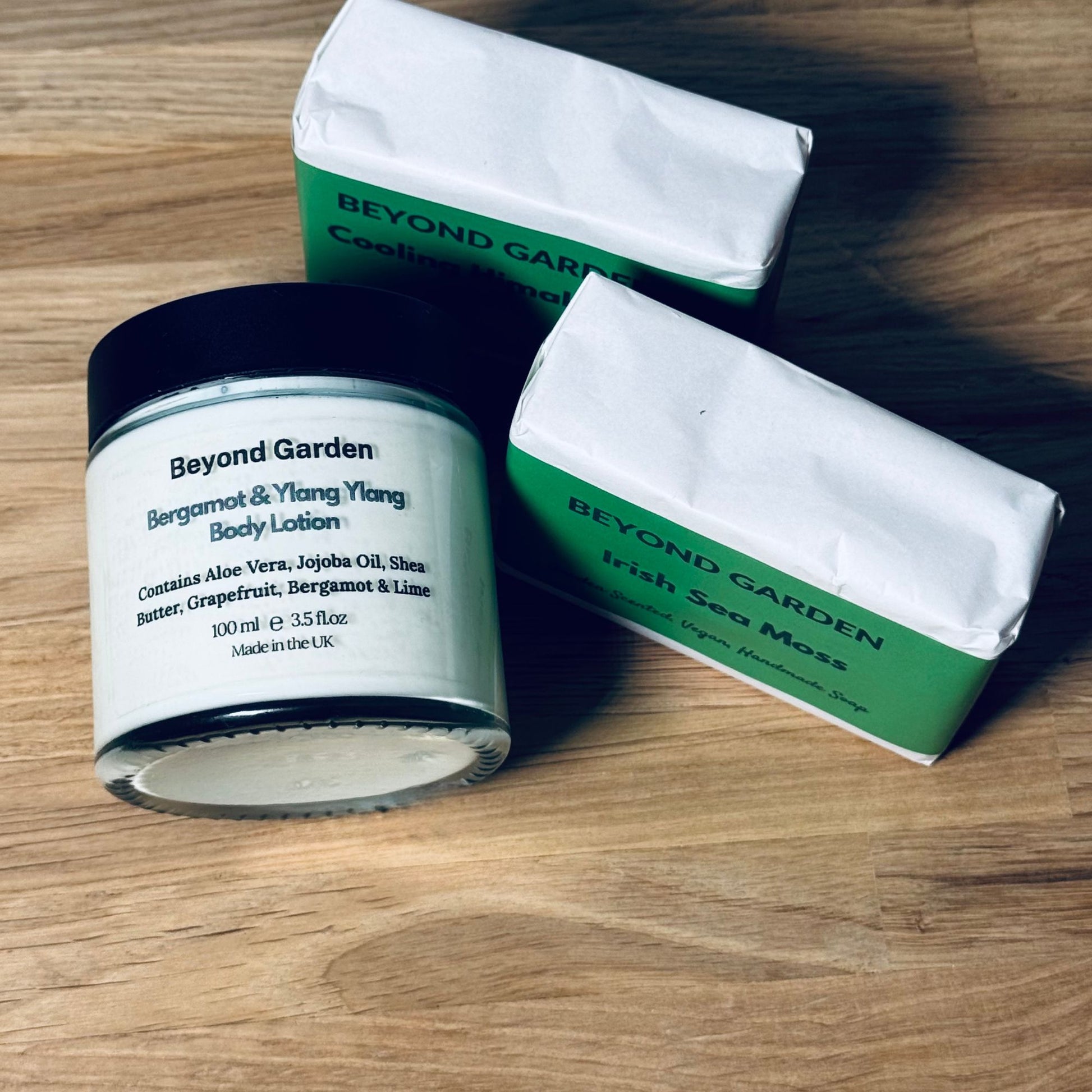 Two bar soaps and a glass jar of lotion lying flat on a table, branded with Beyond Garden labels. The soaps are wrapped in green and white protective paper, with the lotion description visible on the jar.