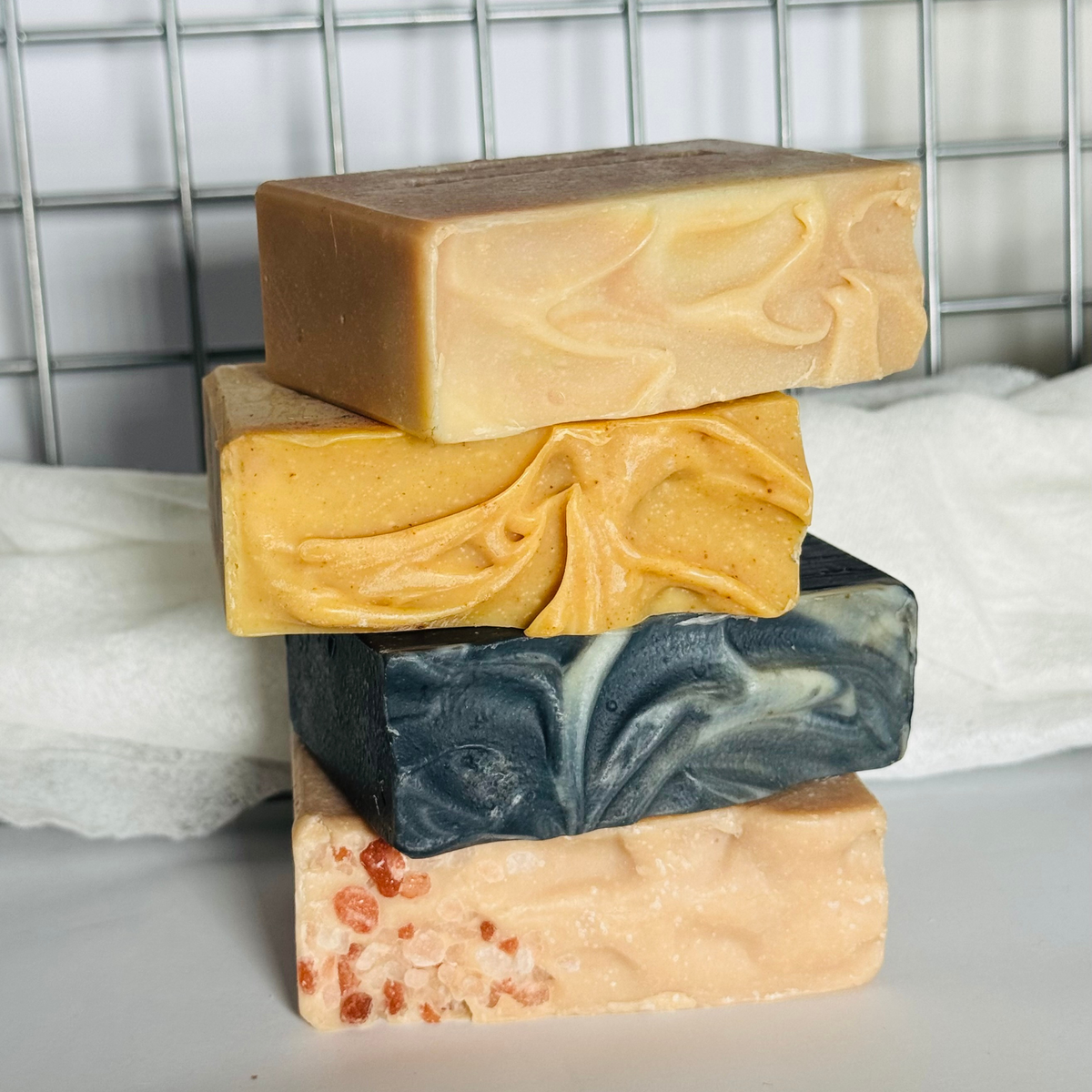 Four handcrafted soaps stacked vertically, showcasing different colours and textures including marbled yellow, dark blue-gray, peach, and speckled with pink salt flecks, set against a background of a white towel and a gray wire rack.