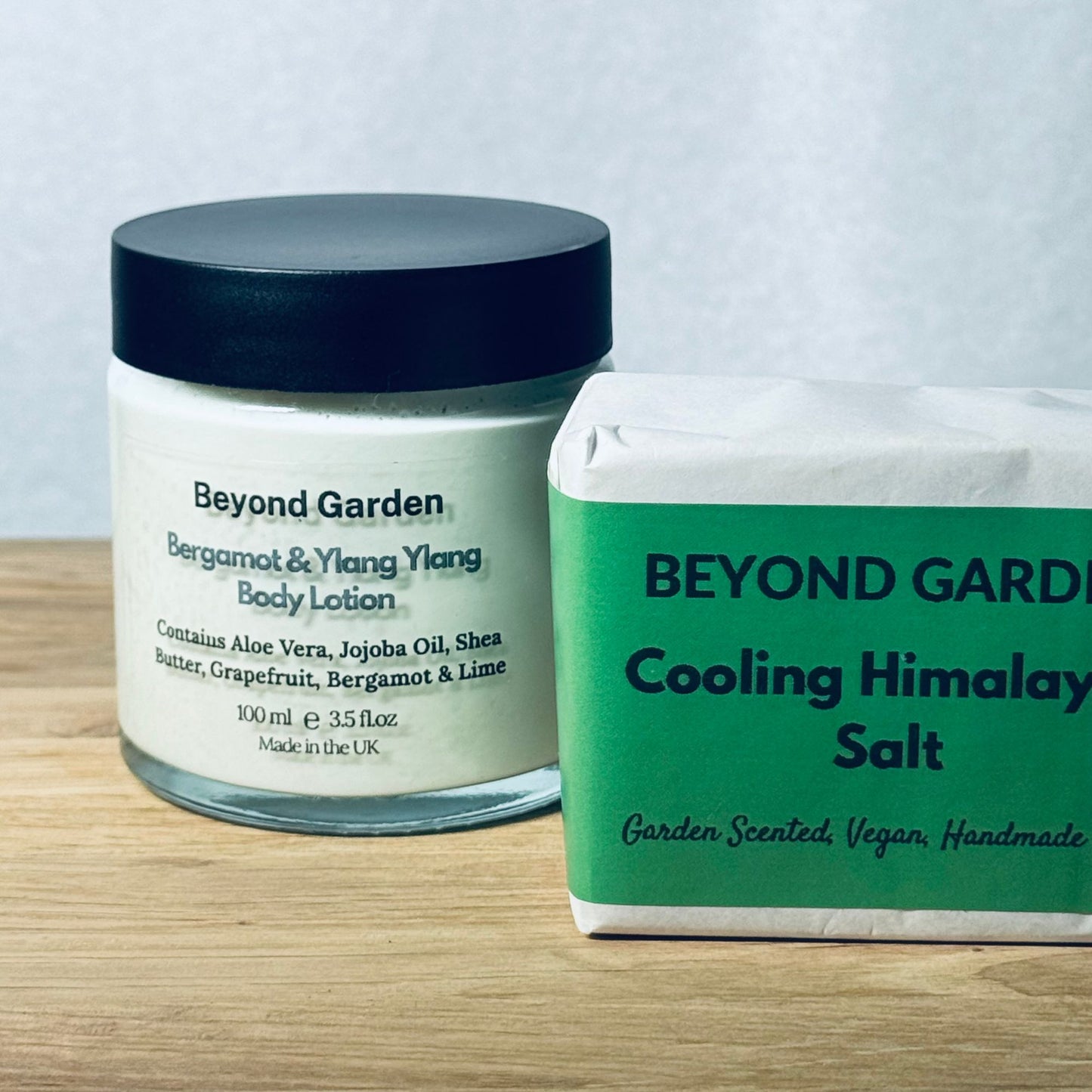 One soap bar branded ‘Cooling Himalayan Salt’ and a glass jar of Bergamot & Ylang Ylang body lotion on a table, both labeled Beyond Garden. The soap is wrapped in beautiful green and white protective paper, with the lotion description visible on the jar. and the soap paper